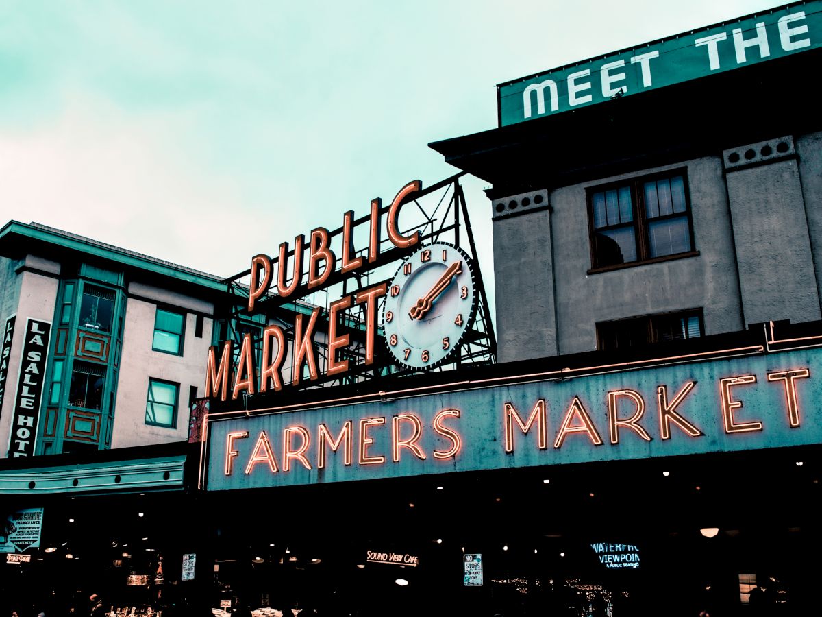 The image shows a neon sign that reads "Public Market" and "Farmers Market," with a clock and surrounding buildings, possibly at Pike Place Market.