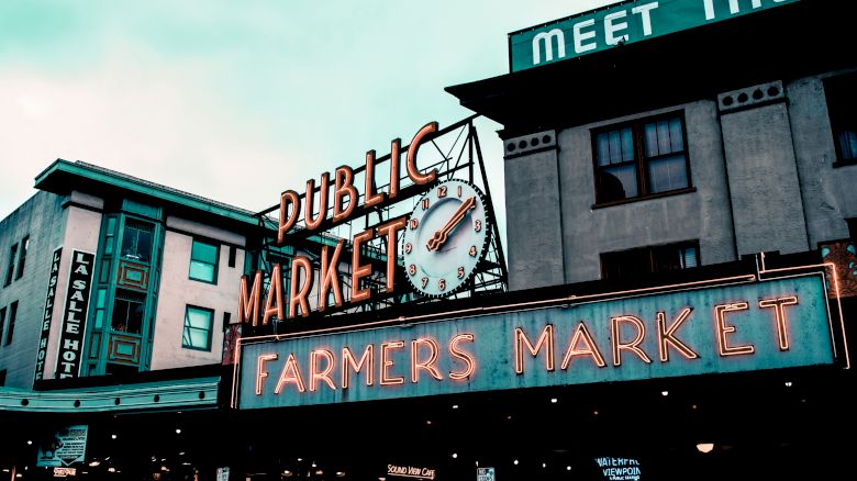 The image shows a neon sign that reads "Public Market" and "Farmers Market," with a clock and surrounding buildings, possibly at Pike Place Market.