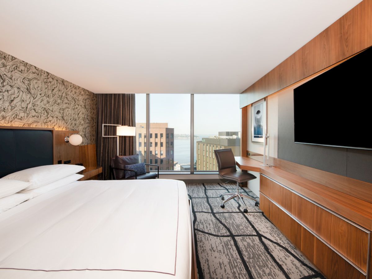 A modern hotel room with a large bed, a wall-mounted TV, a desk, a chair, and a view of city buildings through a large window.