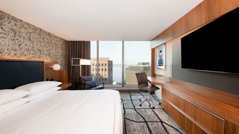 A modern hotel room with a large bed, a wall-mounted TV, a desk, a chair, and a view of city buildings through a large window.