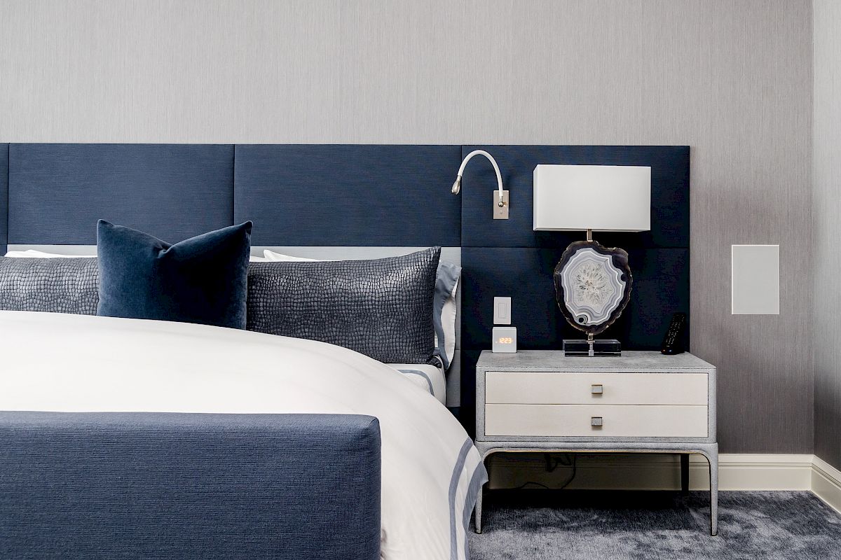A contemporary bedroom with a blue and white color scheme, featuring a bed, nightstand, lamp, and decorative items, next to a soft, carpeted floor.