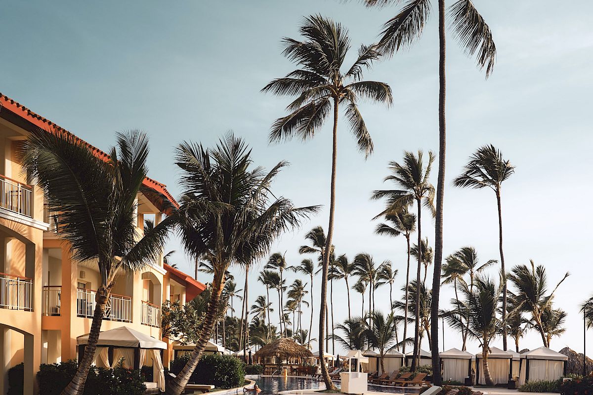 A resort swimming pool area with lounge chairs, surrounded by tall palm trees and an adjacent multi-story building, under a clear sky.