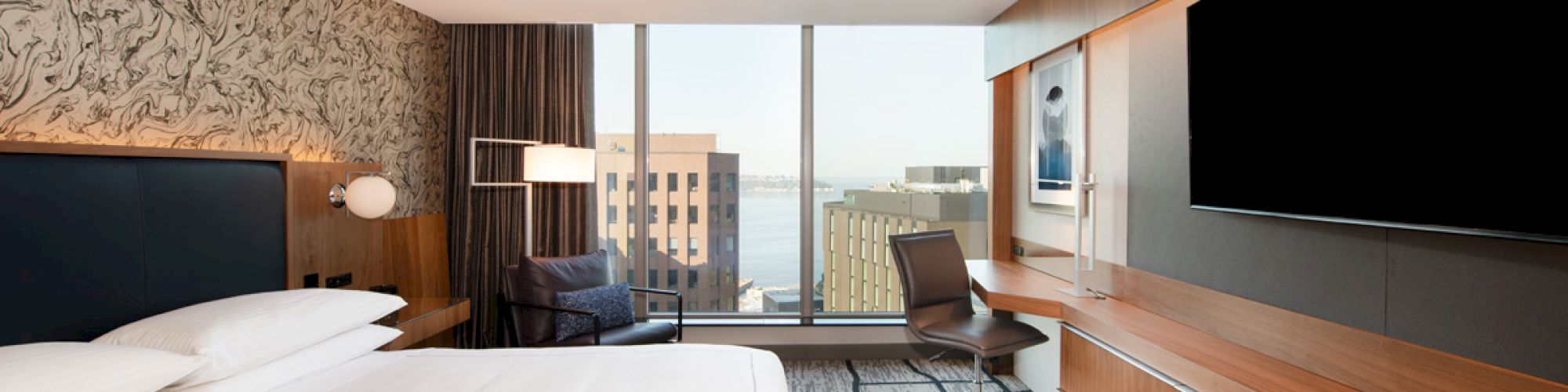 A modern hotel room with a king-size bed, wall-mounted TV, desk with chair, and large windows offering a city view.