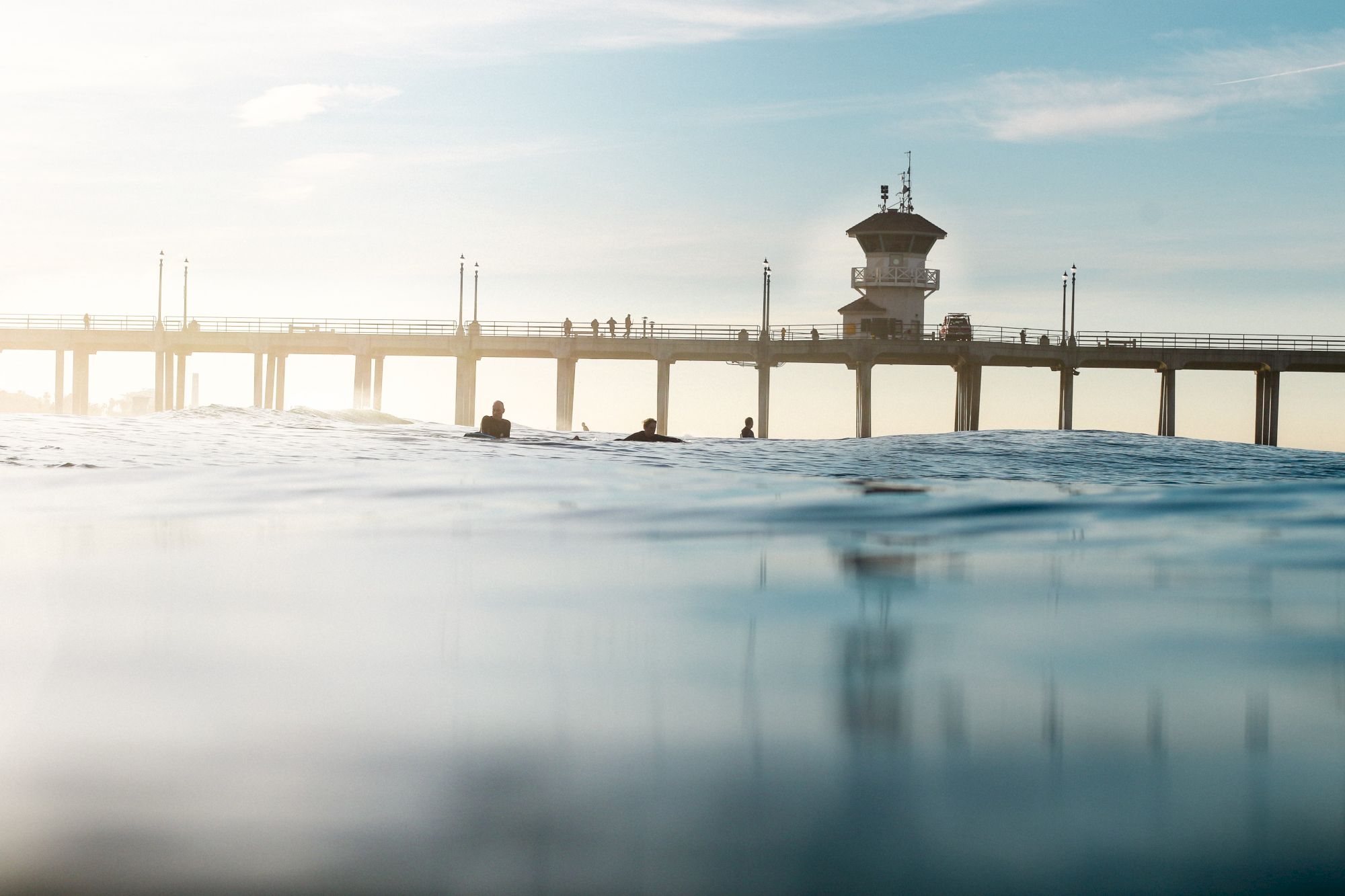 An ocean scene with people in the water near a pier, featuring a structure with a tower and lampposts. The sky is bright and clear.